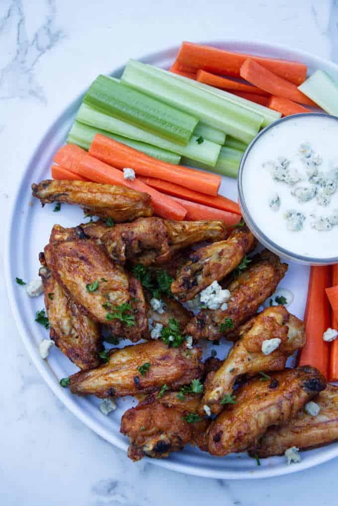 chicken wings with buffalo sauce sitting on a white plate with blue cheese and celery and carrots in the background