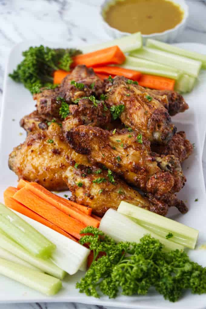 crispy air fried chicken wings sitting on a white plate with sliced carrots and celery with parsley sprinkled on top and dish of honey mustard in background