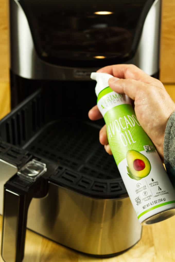 Avocado oil aerosol spray in front of a stainless and black air fryer on a wood table