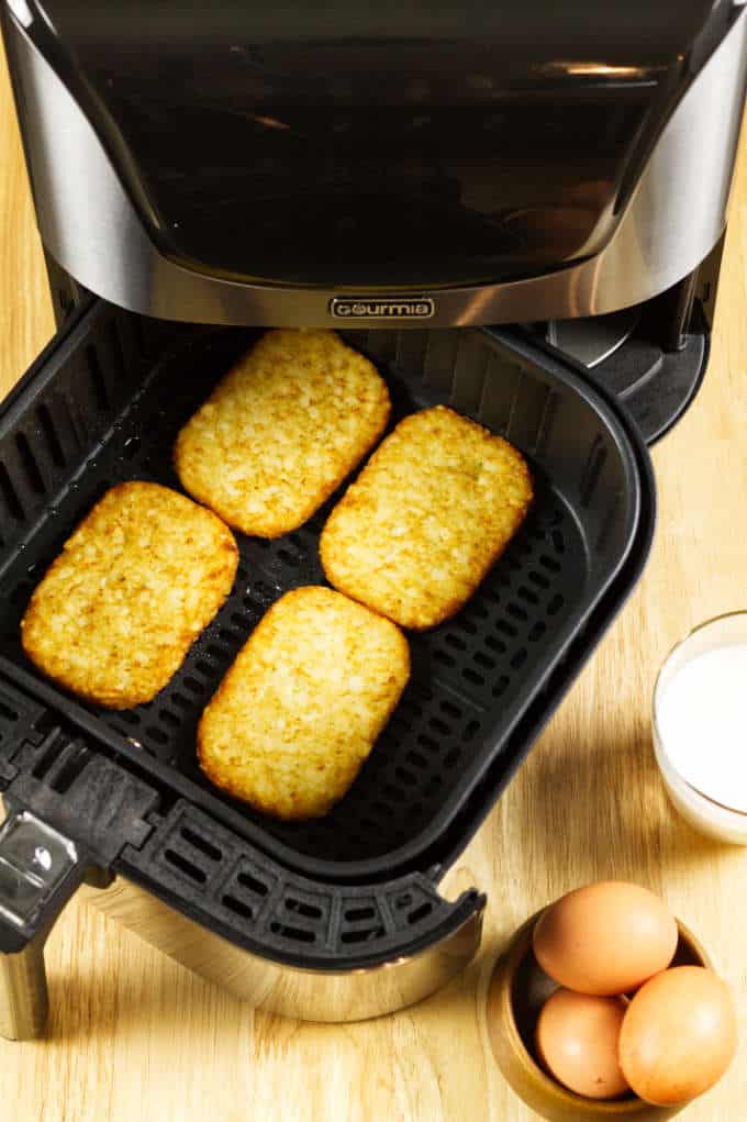 hash browns spaced out in black and stainless air fryer basket with eggs and glass of milk on wood table