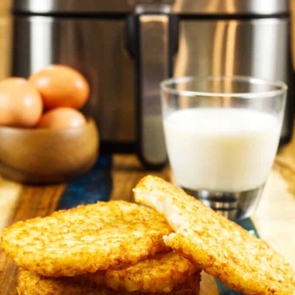 four air fried hash brown patties on wood and blue epoxy cutting board with glass of milk and bowl of brown eggs in back ground and stainless air fryer