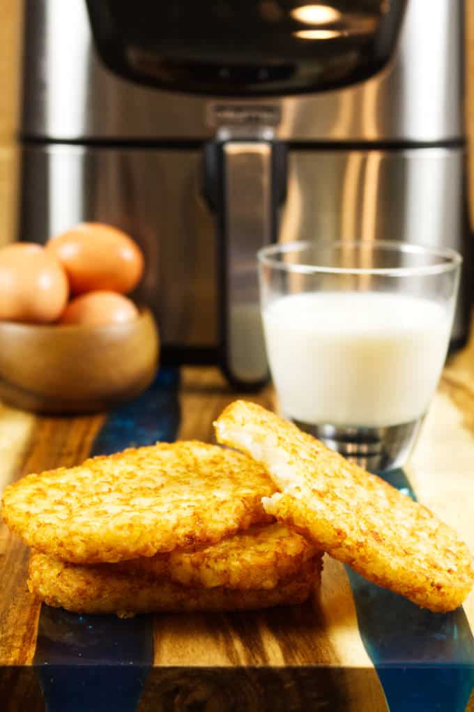 four air fried hash brown patties on wood and blue epoxy cutting board with glass of milk and bowl of brown eggs in back ground and stainless air fryer