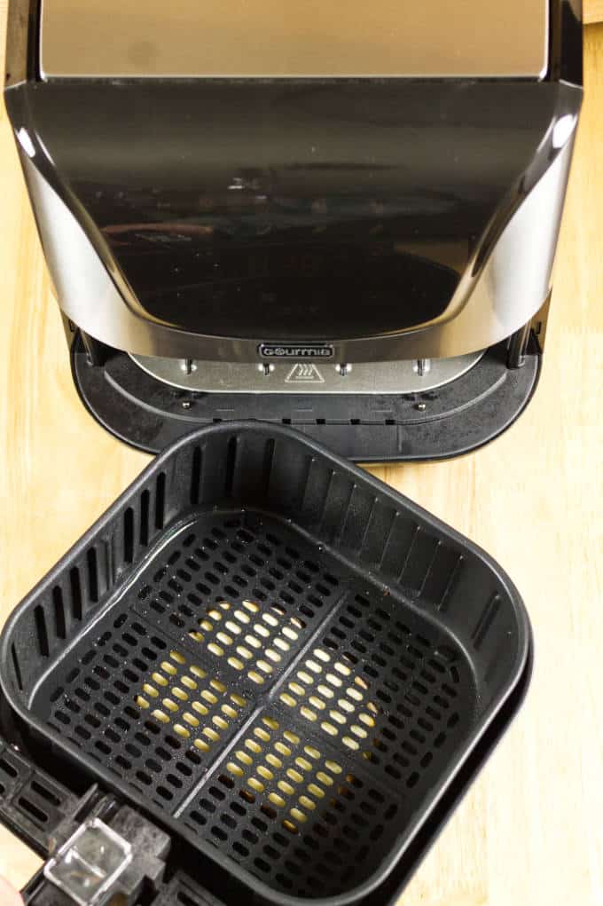 black and stainless air fryer with slice of bread underneath the removable basket and pan on top of wood table