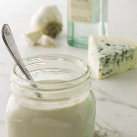 mason jar of filled with blue cheese dressing with spoon sticking out, blue cheese wedge and whole bunch of garlic in the background all sitting on a marble surface