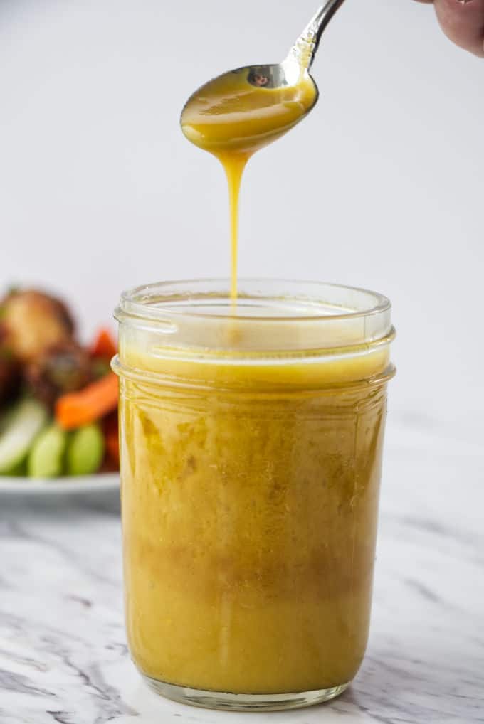brownish yellow honey mustard sauce in mason jar with chicken wings and carrots and celery on a white plate in the background
