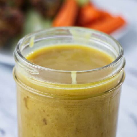 brownish yellow honey mustard sauce in mason jar with chicken wings and carrots and celery on a white plate in the background