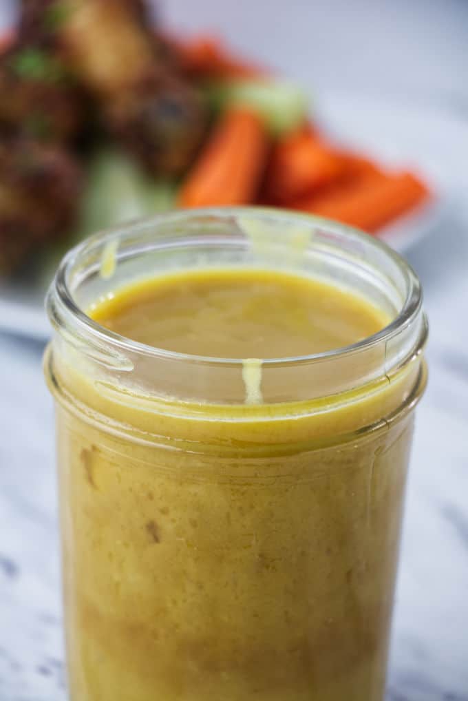 brownish yellow honey mustard sauce in mason jar with chicken wings and carrots and celery on a white plate in the background