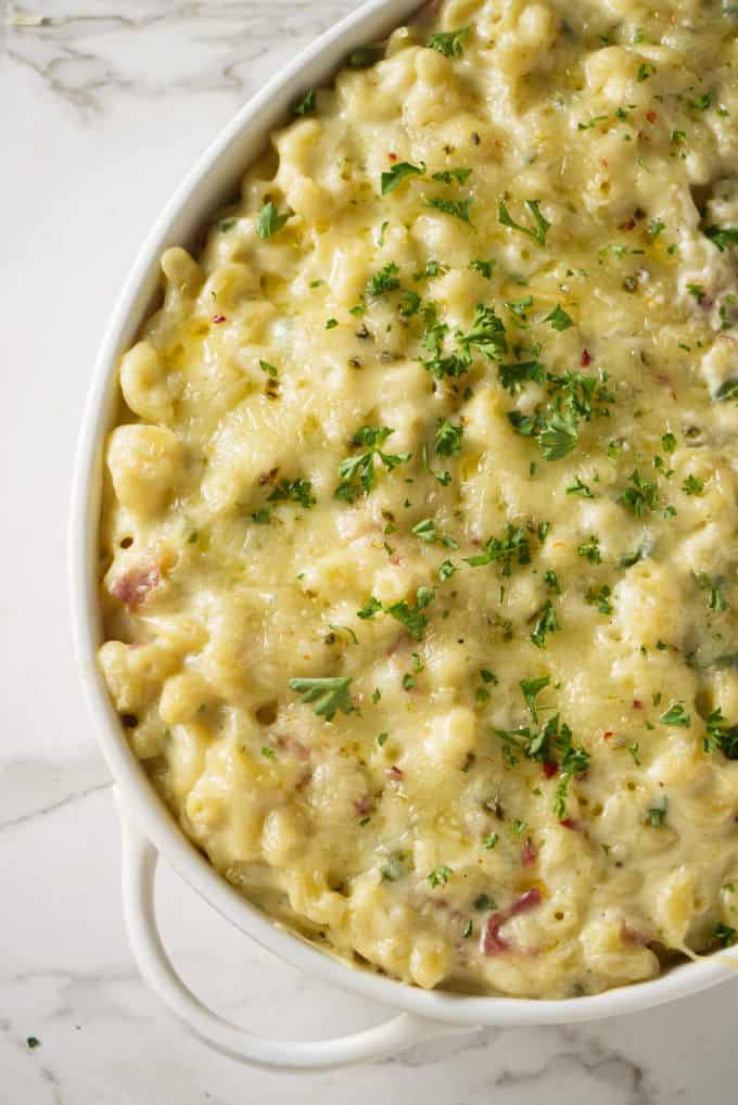 macaroni and cheese in a white ceramic baking dish topped with melted pepper jack cheese with chunks of bacon and topped with green garnish on top of a marble surface