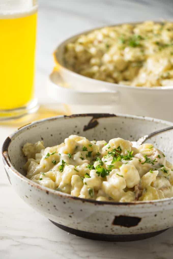 macaroni and cheese in a stone bowl with a spoon and topped with a green garnish. White baking dish in background filled with mac and cheese and a beer in the background