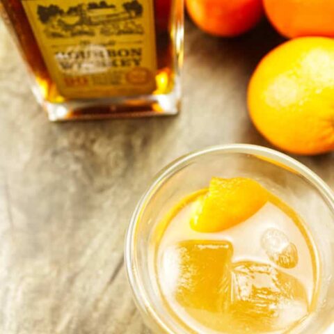 double glass wall insulated rocks glass filled with ice and orange colored mixed cocktail with orange twist inside. Woodinville bourbon and oranges in background sitting on dark stained wood.