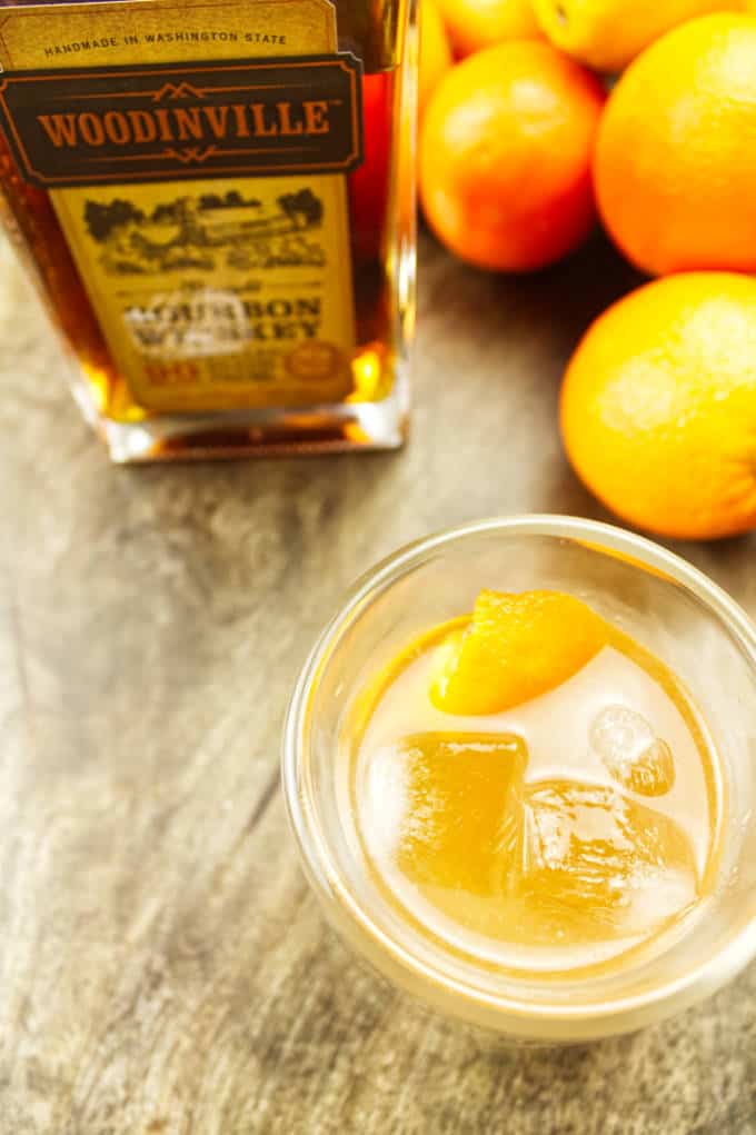 double glass wall insulated rocks glass filled with ice and orange colored mixed cocktail with orange twist inside. Woodinville bourbon and oranges in background sitting on dark stained wood.