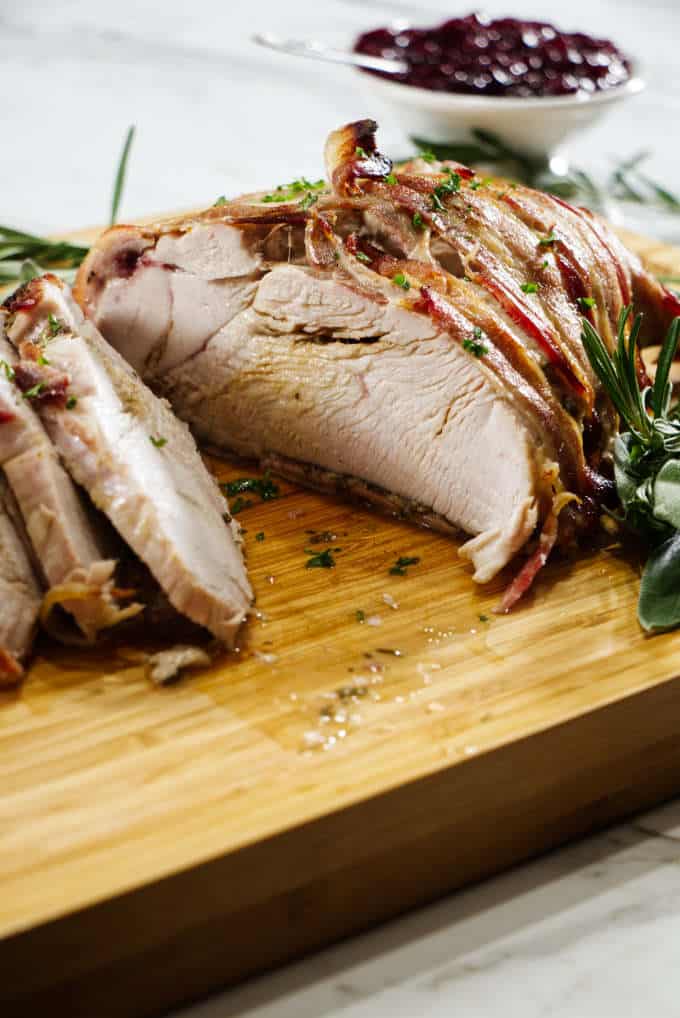 turkey breast wrapped in bacon and sliced sitting on a wooden cutting board with sage in the background with a bowl of cranberry sauce and marble in background