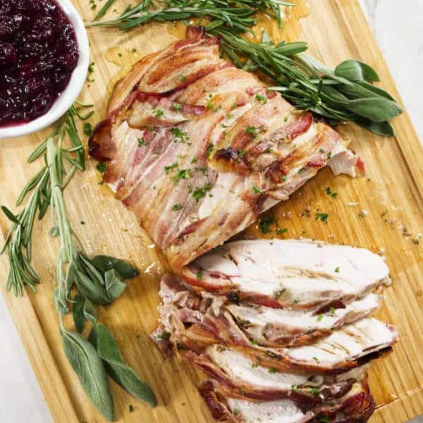 turkey breast wrapped in bacon and sliced sitting on a wooden cutting board with sage in the background with a bowl of cranberry sauce and marble in background