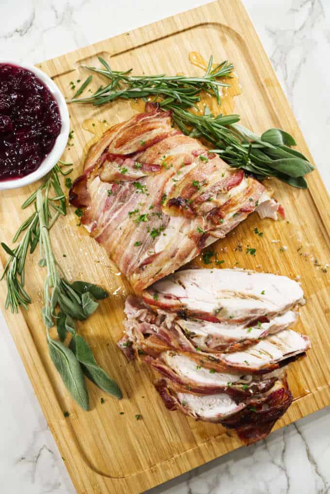 turkey breast wrapped in bacon and sliced sitting on a wooden cutting board with sage in the background with a bowl of cranberry sauce and marble in background