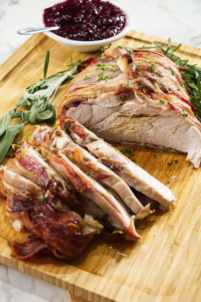 turkey breast wrapped in bacon and sliced sitting on a wooden cutting board with sage in the background with a bowl of cranberry sauce and marble in background