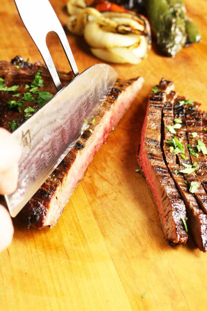 grilled flank steak on wood cutting board being sliced with a chef knife, veggies in background