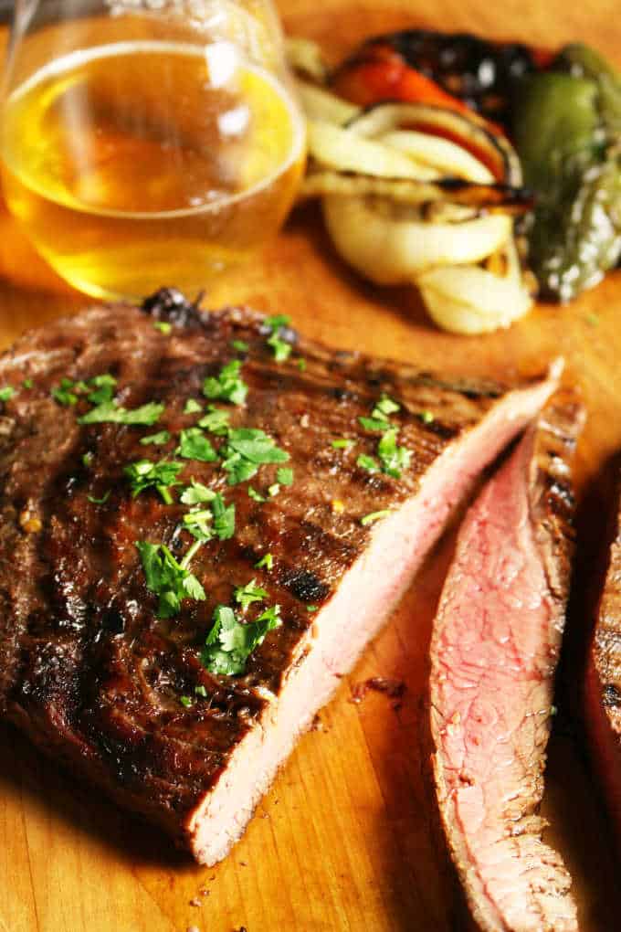 grilled flank steak on wood cutting board sliced into strips medium rare, veggies in background with a glass of beer