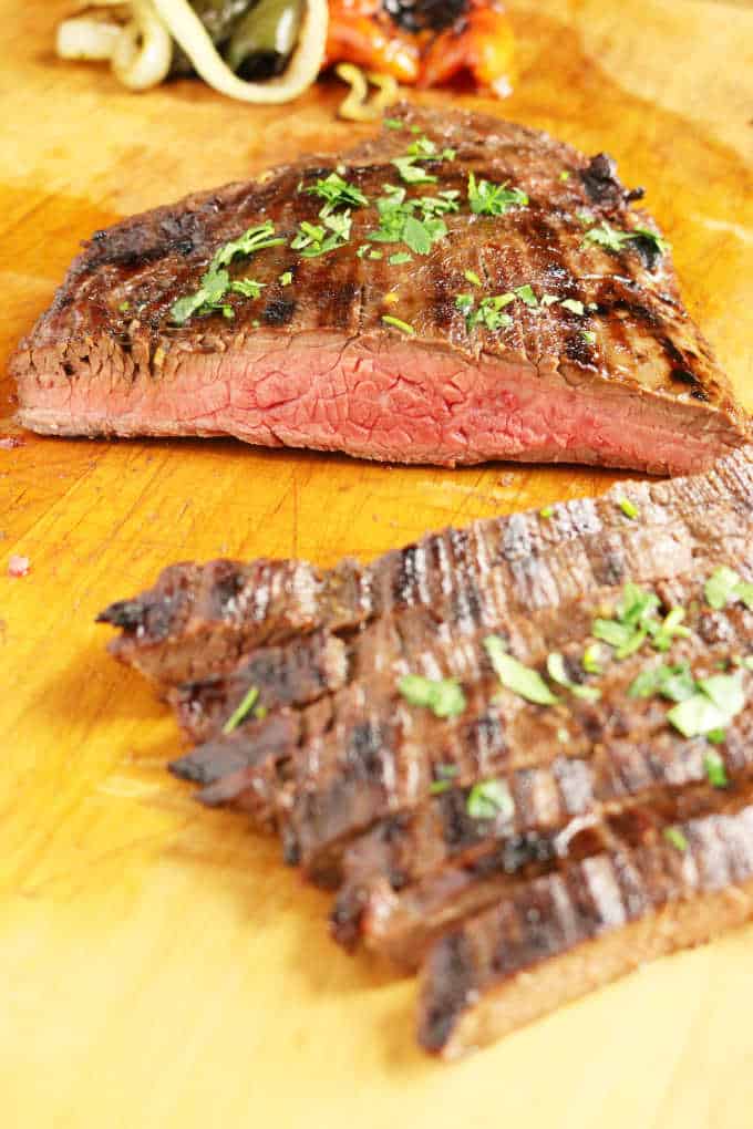 grilled flank steak on wood cutting board sliced into strips medium rare, veggies in background