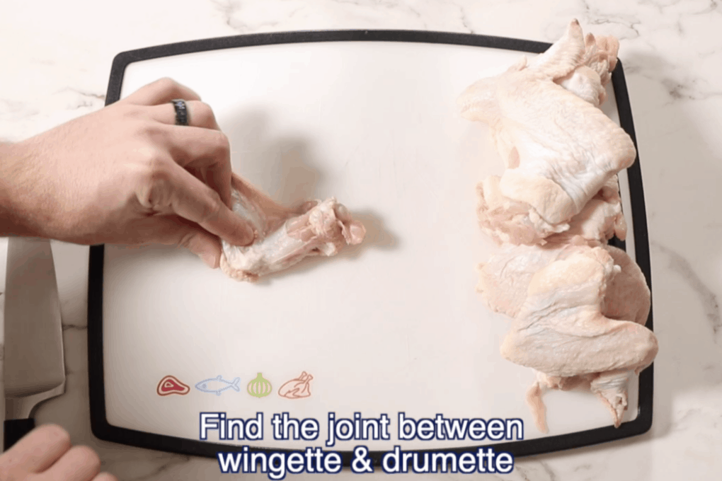 chicken wing on a white cutting board with a hand feeling for the joint between the wingette and drumette