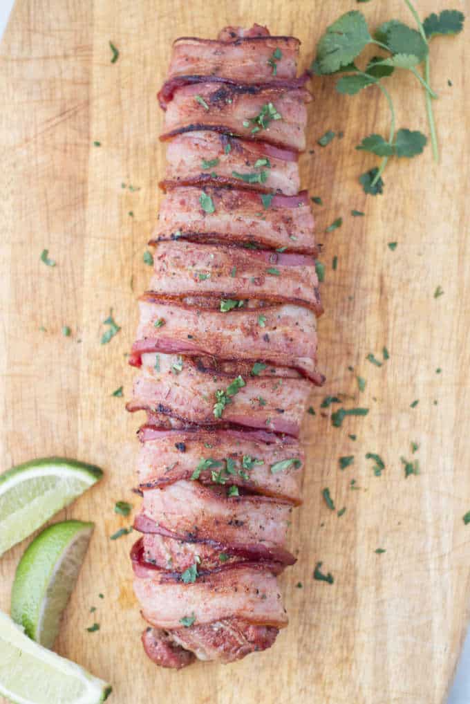 12 slices of bacon wrapped around a pork tenderloin, laying on a wood cutting board with 3 slices of lime and some sprigs of cilantro and chopped cilantro sprinkled on top.