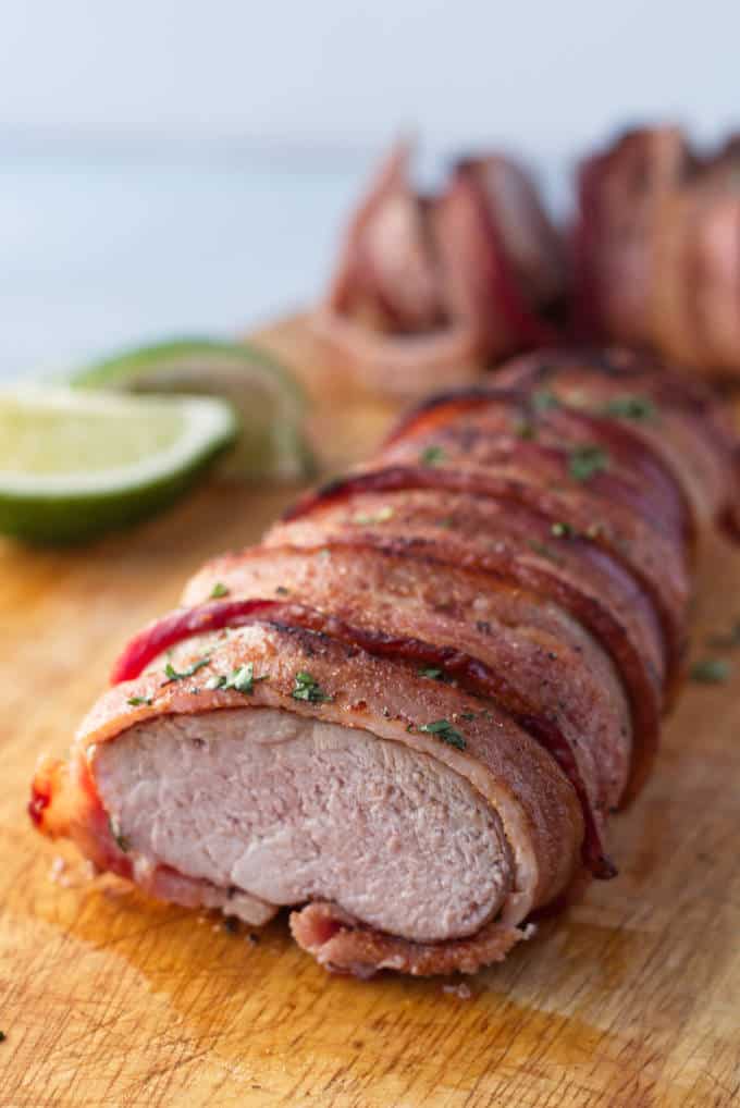 Bacon wrapped pork tenderloin on wood cutting board with cilantro and lime garnish