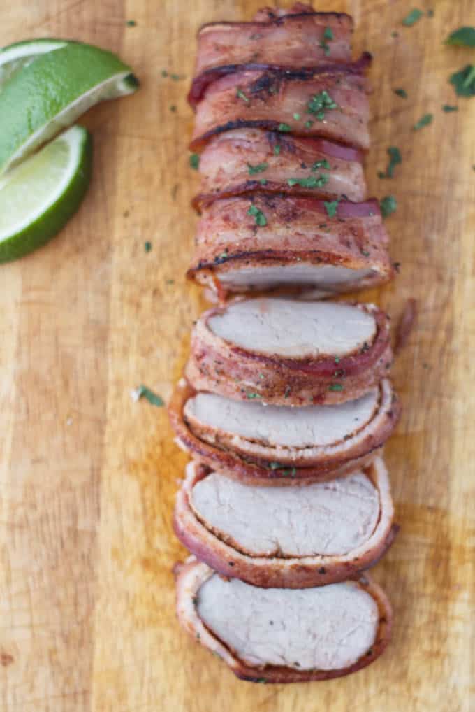 Bacon wrapped pork tenderloin cut into slices on wood cutting board with cilantro and lime garnish