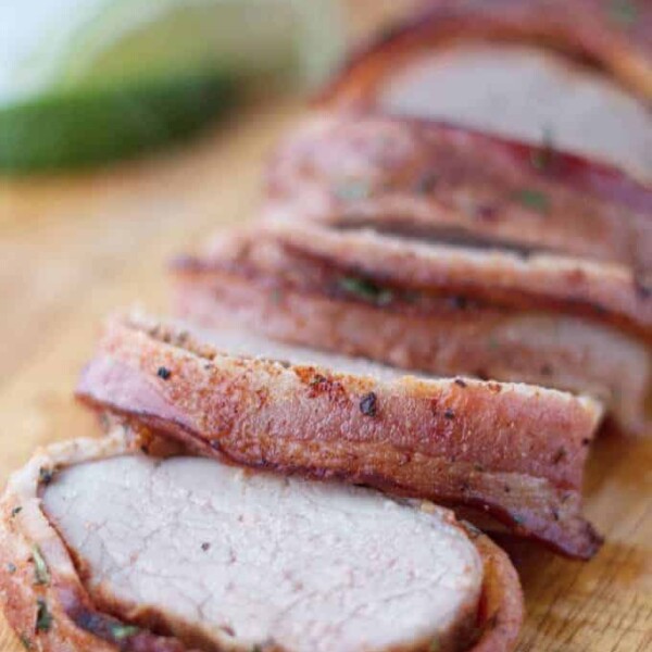 Bacon wrapped pork tenderloin cut into slices on wood cutting board with cilantro and lime garnish