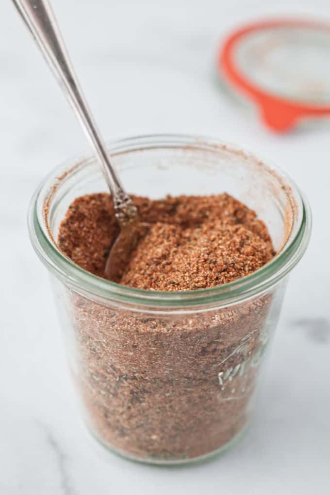 Small glass mason jar filled with brownish red cajun spice seasoning with a silver spoon dug into the spices, mason jar is sitting on a marble table with mason jar lid and orange gasket sitting in background, blurry