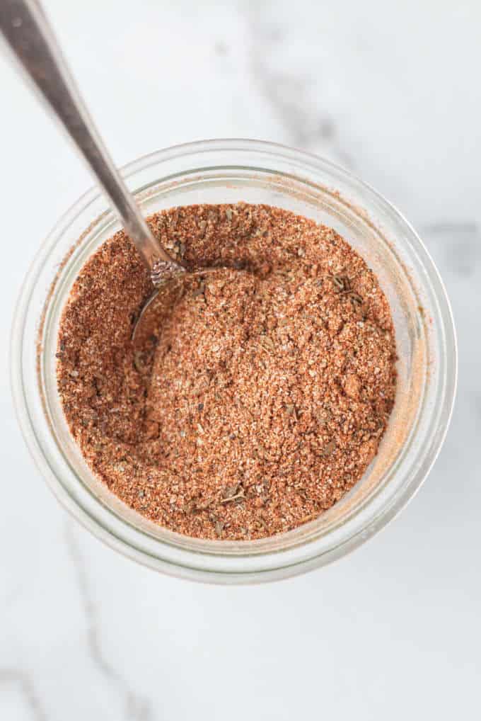 Small glass mason jar filled with brownish red cajun spice seasoning with a silver spoon dug into the spices, mason jar is sitting on a marble table