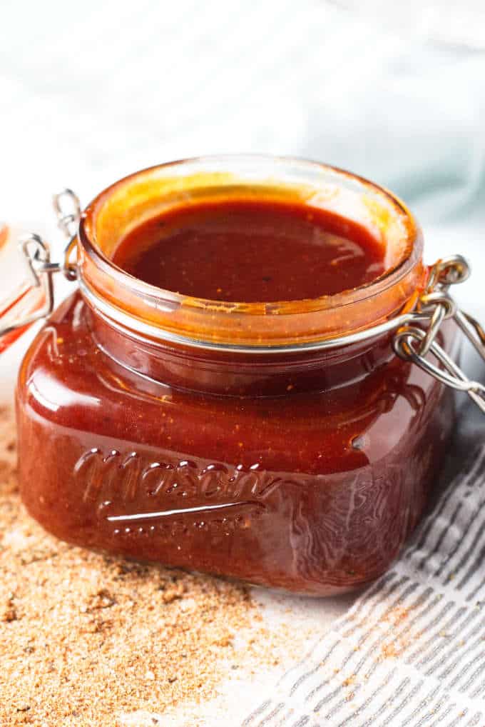 Kansas City-style BBQ sauce in a mason jar sitting on gray and white napkin with some loose seasoning on the table in front