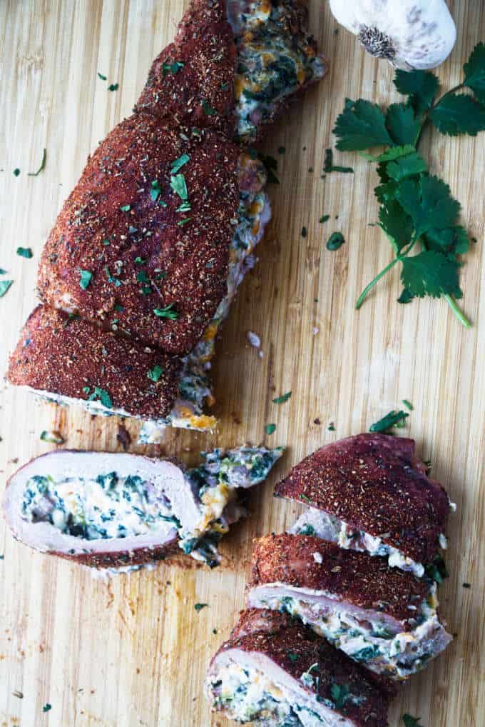 sliced stuffed pork tenderloin with cajun rub and twine holding it together, cheese stuffing, cilantro and garlic cloves sitting on wood