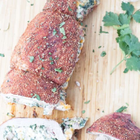 sliced stuffed pork tenderloin with cajun rub and twine holding it together, cheese stuffing, cilantro and garlic cloves sitting on wood