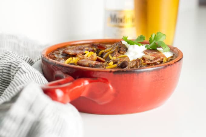 small red pot filled with brisket chili with cheese and sour cream and a garnish of cilantro, modelo beer bottle with glass of beer in background and napkins in foreground