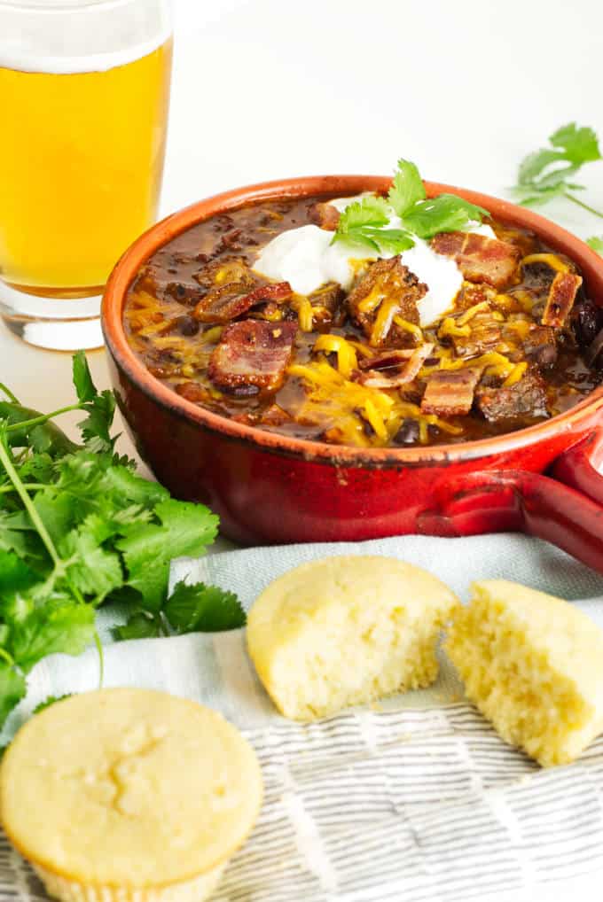 small red pot filled with brisket chili with cheese and sour cream and a garnish of cilantro, glass of beer in the background and cornbread in the foreground