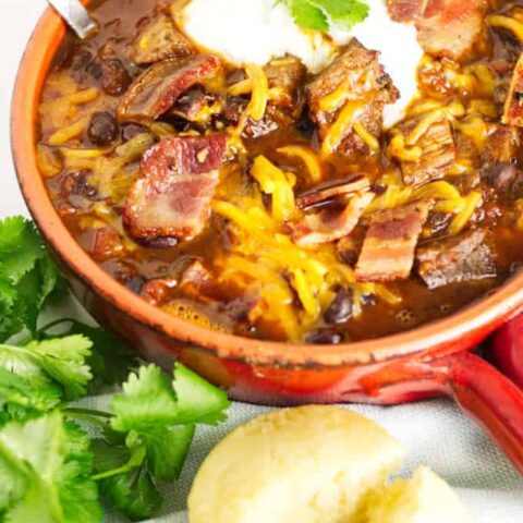 small red pot filled with brisket chili with cheese and sour cream and a garnish of cilantro, cornbread muffin in the foreground