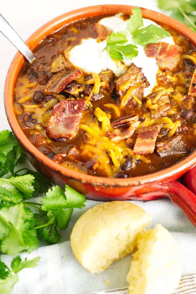 small red pot filled with brisket chili with cheese and sour cream and a garnish of cilantro, cornbread muffin in the foreground