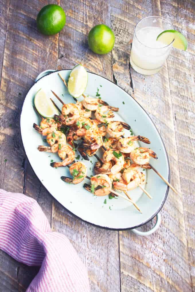 Grilled tequila lime shrimp skewers on a blue platter with limes and a margartia