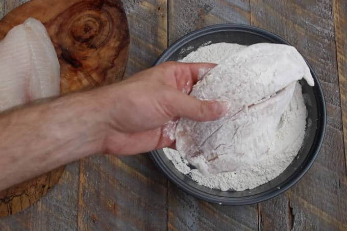 tilapia filet being coated in flour in baking pan