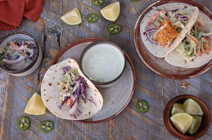 two plates on wooden table with tortillas, tilapia fish tacos being built, bed of slaw on each tortilla and chunk of tortilla in one