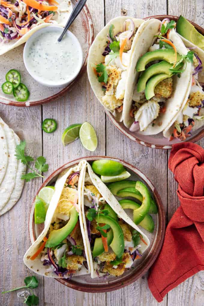 Two plates of tilapia fish tacos with avocados and lime slices.