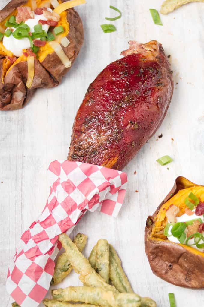 Smoked wild turkey leg laying on white cutting board with loaded baked sweet potatoes and some fried green beans