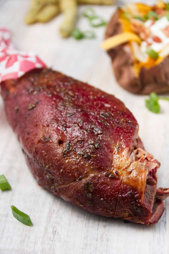 Smoked wild turkey leg laying on white cutting board with loaded baked sweet potatoes and some fried green beans