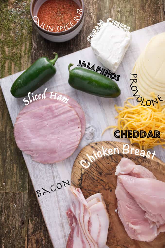Ingredients for smoked jalapeño popper chicken: cajun spice rub, cream cheese, jalapeños, sliced ham, provolone cheese, cheddar cheese, bacon, and chicken breast.