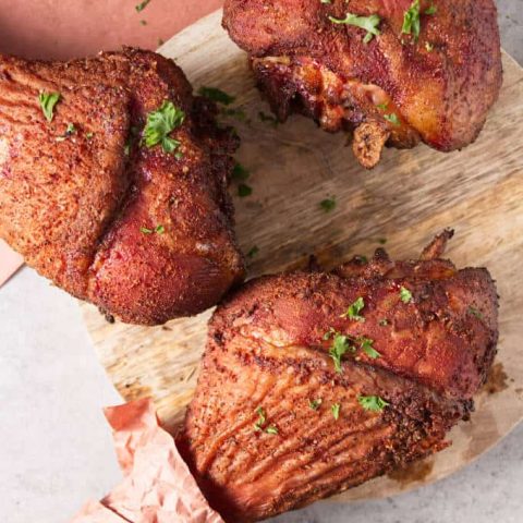 Three smoked turkey legs laying on a concrete table and circular wood cutting board with peach butcher paper