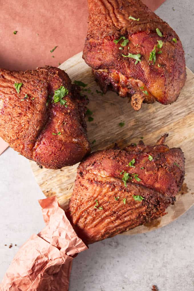 Three smoked turkey legs laying on a concrete table and circular wood cutting board with peach butcher paper