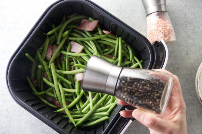 Adding pepper to an air fryer with green beans and bacon.