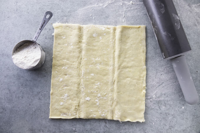 A sheet of puff pastry next to a rolling pin and some flour.
