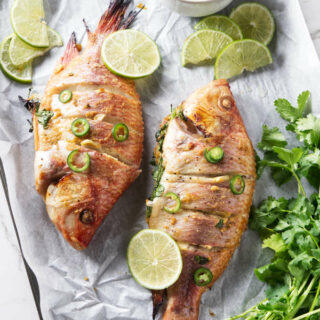 Grilled tilapia on a tray with a bowl of sauce, lime slices, and herbs.