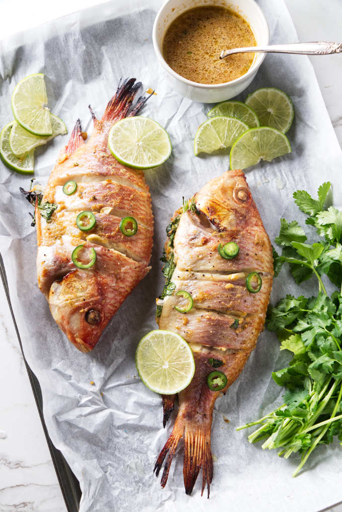 Grilled tilapia on a tray with a bowl of sauce, lime slices, and herbs.