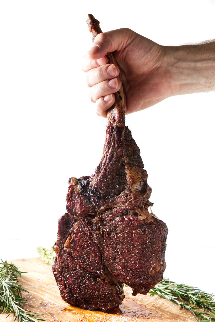 seared tomahawk steak being held vertically from the bone, resting on cutting board